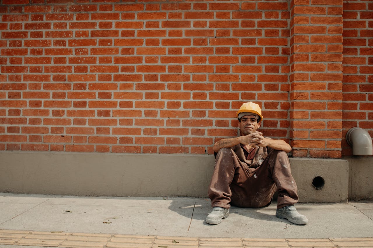 ¿Y si pedir ayuda es lo más valioso que puedes hacer hoy? Tired construction worker sitting against brick wall in sunny urban setting.